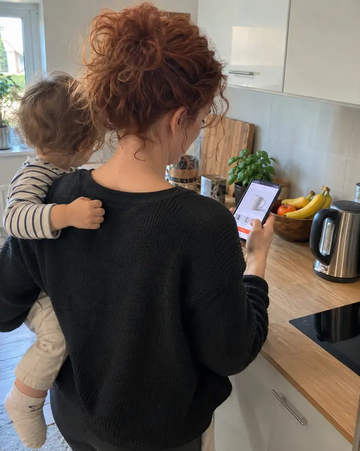 Frau mit Kleinkind auf der Hüfte bestellt in der Küche mit einer Hand am Handy