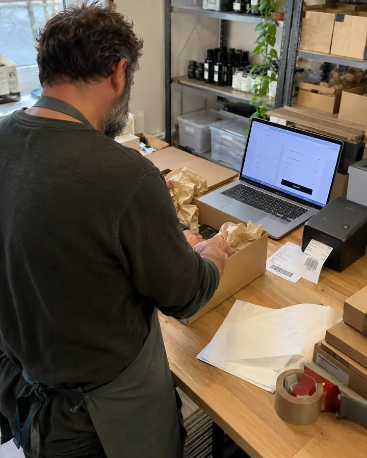 Händler packt eine Bestellung, daneben der Laptop mit der Kasse des Onlineshops und ein Etikettendrucker